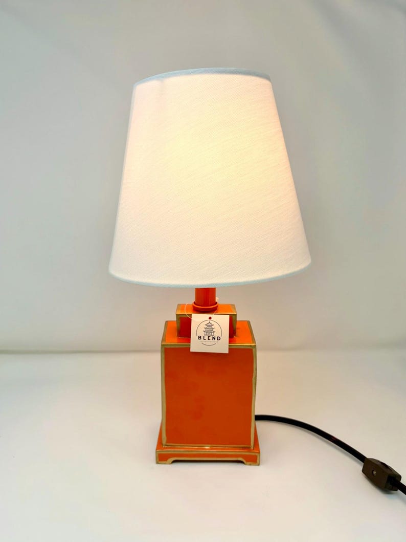 May include: An orange table lamp with a white lampshade and a light blue trim. The lamp has a rectangular base with gold trim and a matching orange stem. A tag with the text "BLEND" is attached to the lamp.