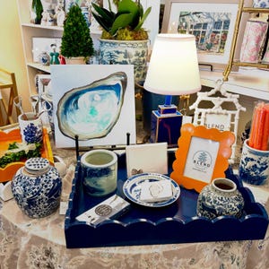 May include: A display of decorative items, including a blue scalloped tray, an orange picture frame, and a blue and white porcelain jar. An oyster painting and a small lamp are also featured. The items are arranged on a patterned tablecloth.