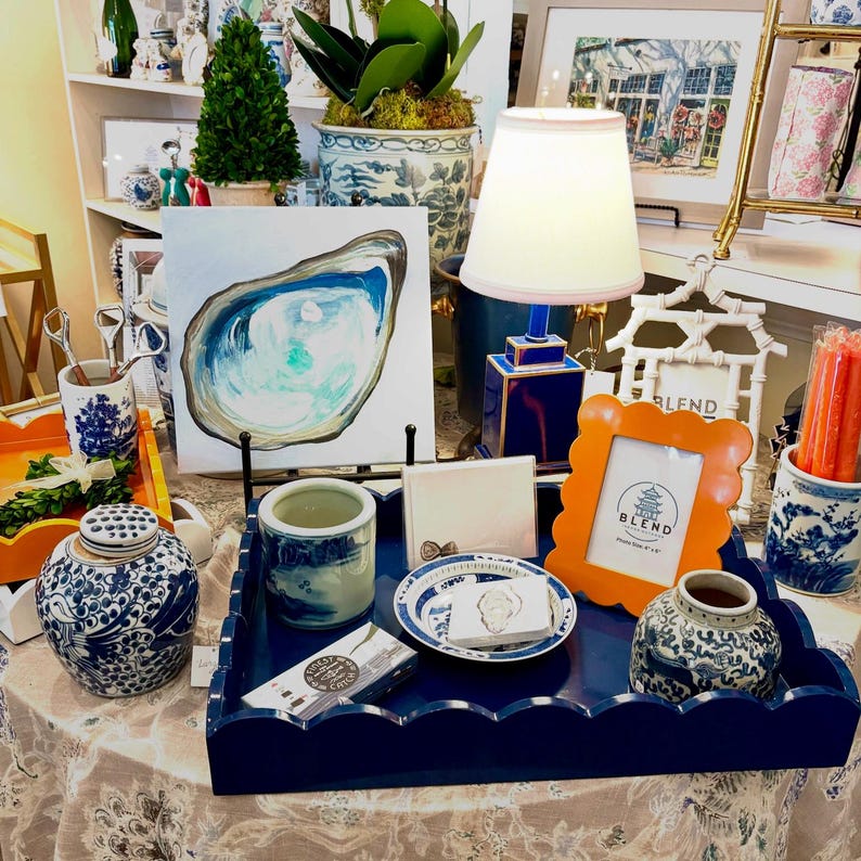 May include: A display of decorative items, including a blue scalloped tray, an orange picture frame, and a blue and white porcelain jar. An oyster painting and a small lamp are also featured. The items are arranged on a patterned tablecloth.