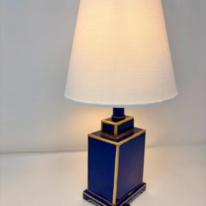 May include: A blue table lamp with a white lampshade. The lamp base is a rectangular shape with gold trim. The lampshade is a light cream color with a light blue trim. A black power cord extends from the base.