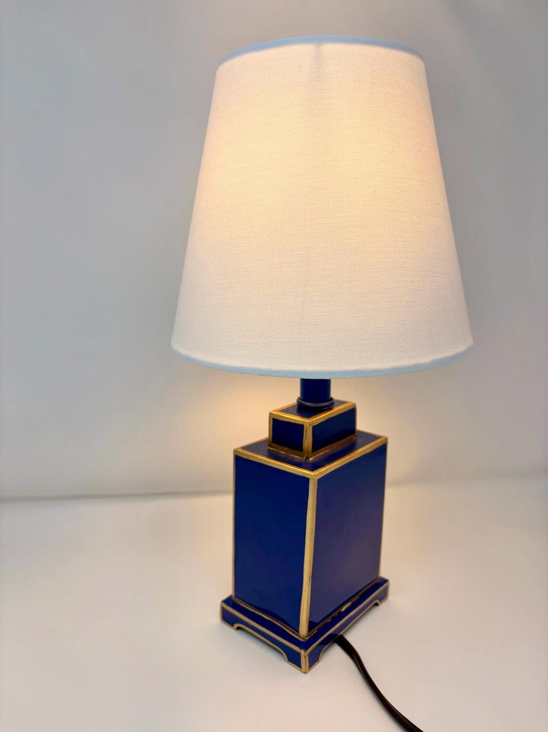 May include: A blue table lamp with a white lampshade. The lamp base is a rectangular shape with gold trim. The lampshade is a light cream color with a light blue trim. A black power cord extends from the base.