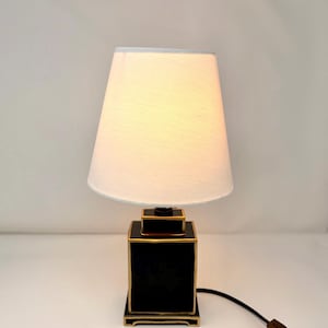 May include: A black and gold table lamp with a white lampshade. The lamp has a square base with gold trim and a matching top. The lampshade is a classic tapered shape, and the lamp is plugged into an outlet.