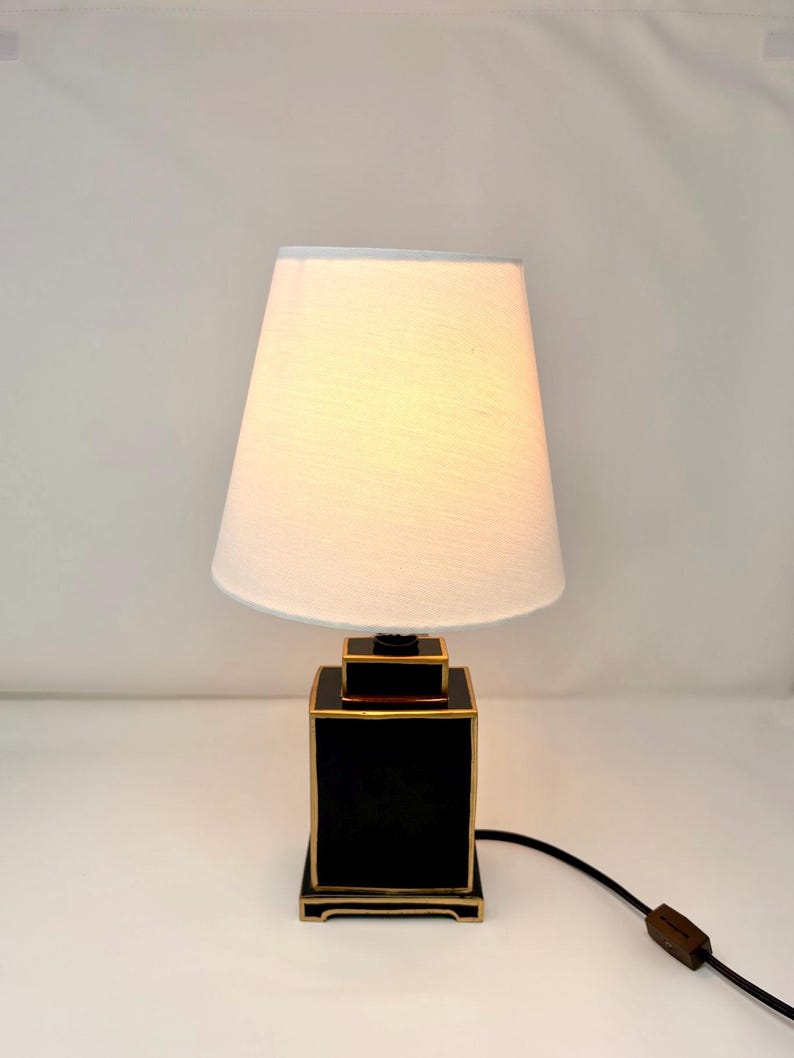 May include: A black and gold table lamp with a white lampshade. The lamp has a square base with gold trim and a matching top. The lampshade is a classic tapered shape, and the lamp is plugged into an outlet.
