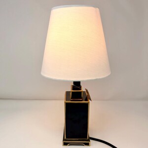 May include: A black and gold table lamp with a white lampshade. The lamp has a rectangular base with gold trim and a black body. The lampshade is a light cream color and tapers towards the top. A black power cord extends from the base.