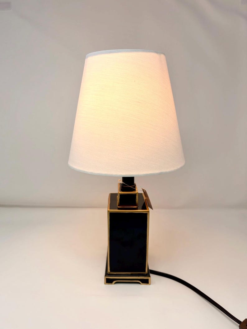 May include: A black and gold table lamp with a white lampshade. The lamp has a rectangular base with gold trim and a black body. The lampshade is a light cream color and tapers towards the top. A black power cord extends from the base.