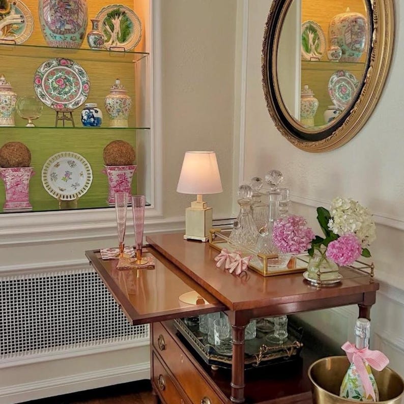 May include: A wooden bar cart with a drop-leaf top, holding a lamp, decanters, and a floral arrangement. A champagne bottle sits in a gold ice bucket. Decorative plates and vases are displayed in a glass cabinet in the background.