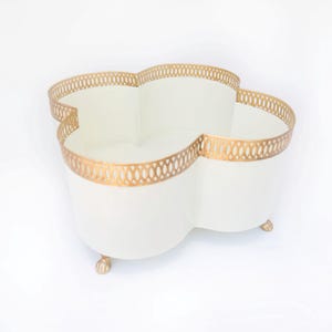 May include: White metal planter with a gold decorative trim and four legs. The planter is shaped like a four-leaf clover.