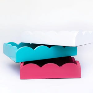 May include: Three decorative trays stacked on top of each other. The top tray is white, the middle tray is turquoise blue, and the bottom tray is pink. All three trays have a scalloped edge.