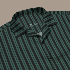 以下が含まれることがあります： 緑と黒のストライプのボタンダウンシャツ。シャツは縦縞模様で、襟が付いています。「Happily Ever After Hours」の文字が襟の内側に表示されています。シャツはクラシックでカジュアルなスタイルです。