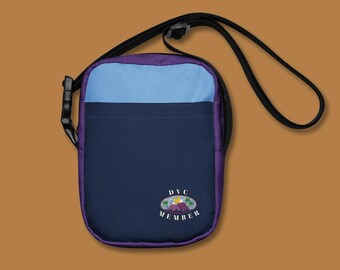 May include: A rectangular crossbody bag with a black strap. The bag is divided into three sections: a light blue top, a navy blue centre, and a purple border. The bag features a small embroidered design with the text "DVC MEMBER".