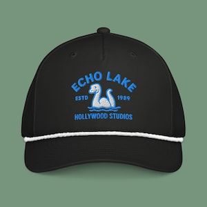 May include: Black baseball cap with a white rope detail and a curved brim. The cap features blue embroidered text that reads "ECHO LAKE" above a cartoon lake monster and "HOLLYWOOD STUDIOS" below. The text also includes "ESTD 1989".
