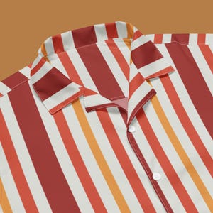 Può includere: Una camicia abbottonata a righe dai colori vivaci. La camicia presenta strisce verticali nelle tonalità del rosso, arancione e bianco. Il colletto e i bottoni sono visibili, mettendo in risalto il design della camicia. Tessuto leggero.