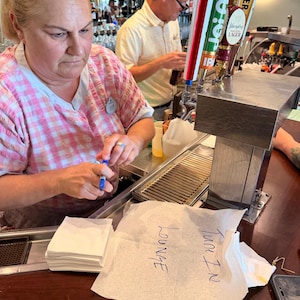 Puede incluir: Una mujer con una camisa a cuadros rosa y blanca est&aacute; de pie detr&aacute;s de una barra, sosteniendo un bol&iacute;grafo. Un hombre con una camisa amarilla est&aacute; detr&aacute;s de ella. Se ven grifos de cerveza. Una servilleta dice "LOUNGE" y "TURN IN".
