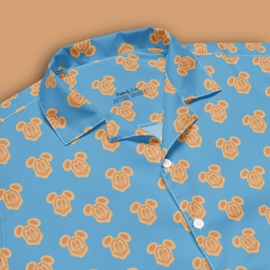May include: Light blue button-down shirt featuring a repeating pattern of orange Mickey Mouse-shaped waffles. The shirt has a collar and white buttons. The text "Happily Ever After Hours" is printed on the collar.