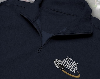 May include: A navy blue quarter-zip pullover featuring an embroidered logo on the chest. The logo depicts a white monorail train with the text BAY LAKE TOWER in white capital letters, underlined by a curved gold swoosh graphic element.