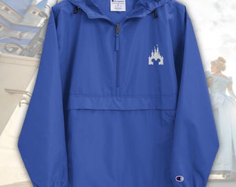 May include: Royal blue Champion anorak jacket with a half-zip and a hood. The jacket has a white castle graphic on the front and the Champion logo on the sleeve. The background includes a blurred image of a castle and a woman in a white dress.