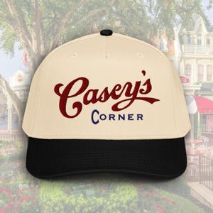 May include: A baseball cap with a cream-colored crown and a black brim. The cap features the text "Casey's Corner" in maroon script and blue block letters. The cap has a black button on top.