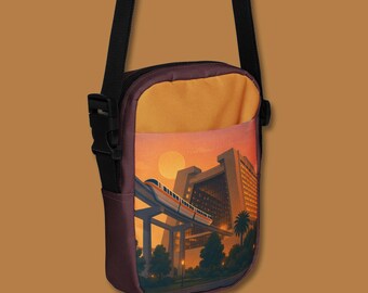May include: A crossbody bag with a sunset scene. The bag has a vibrant orange and purple colour scheme, with a detailed illustration of a monorail and a large building. The bag has a black strap and a black buckle.
