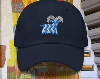 May include: A navy blue baseball cap with an embroidered blue and white goat design. The goat has large horns and a zig-zag pattern on its body.