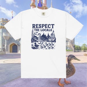 Może przedstawiać: Biała koszulka z niebieskim nadrukiem przedstawiającym kaczkę i kaczątka idące przed zamkiem. Tekst "Respect the Locals" znajduje się nad nadrukiem.
