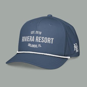 May include: A navy blue baseball cap with white text that reads "EST. 2019 RIVIERA RESORT ORLANDO, FL." A white rope detail accents the brim. The cap has a curved brim and a small logo on the side.