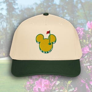 Könnte beinhalten: Cremefarbene Baseballkappe mit dunkelgrünem Schirm und Knopf. Die Kappe hat ein gelb-grünes gesticktes Design eines Golfplatzes mit roter Fahne, geformt wie Mickey Mouse Ohren.