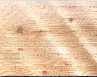 Op de afbeelding: Een houten Amerikaanse vlag met de Pledge of Allegiance erin gegraveerd. Het vlagontwerp bevat sterren en strepen, met de tekst gebeiteld in een schreeflettertype. Het hout heeft een natuurlijke, lichtbruine kleur.