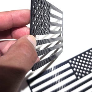 Könnte beinhalten: Schwarz-weißes Design der amerikanischen Flagge. Die Flagge besteht aus einem dunklen Material mit gewebter Textur, wobei Sterne und Streifen ausgeschnitten sind. Eine weitere Flagge liegt flach im Hintergrund.