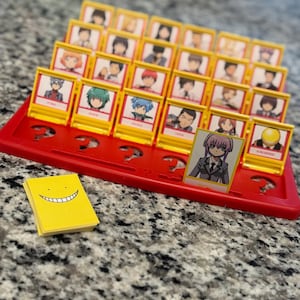 May include: A red Guess Who? board game with yellow character cards featuring anime-style illustrations. A small stack of yellow cards with a smiling face is on the side. The game is set on a speckled grey surface.