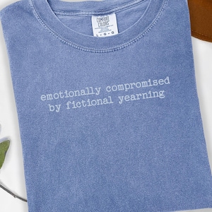 Puede incluir: Una camiseta azul Comfort Colors con la frase "emotionally compromised by fictional yearning" en letras blancas. Un sombrero marrón es parcialmente visible en el fondo. La camiseta está doblada y sobre una superficie blanca.