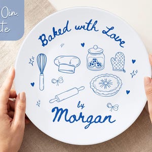 May include: A white ceramic plate, 10 inches in diameter, with blue illustrations of baking tools and the words "Baked with Love by Morgan." The plate features a whisk, chef's hat, jar, oven mitt, pie, and rolling pin.