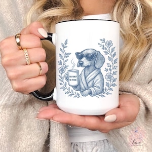 May include: White ceramic mug with a black handle and rim. The mug features a blue illustration of a dachshund wearing sunglasses and a robe, holding a coffee cup. The cup reads "Too Fancy to Fetch". The design is surrounded by blue floral elements.