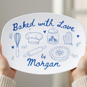 May include: White oval serving platter with blue illustrations of baking tools and the text "Baked with Love by Morgan." The design includes a whisk, chef's hat, rolling pin, pie, and oven mitt.