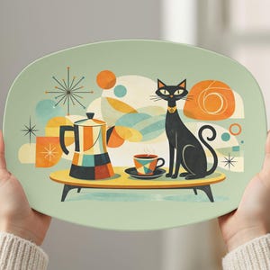 May include: A decorative serving platter with a mid-century modern design. The platter features a black cat, coffee pot, and cup on a yellow table. The background has orange, blue, and white geometric shapes. The platter is held by two hands.