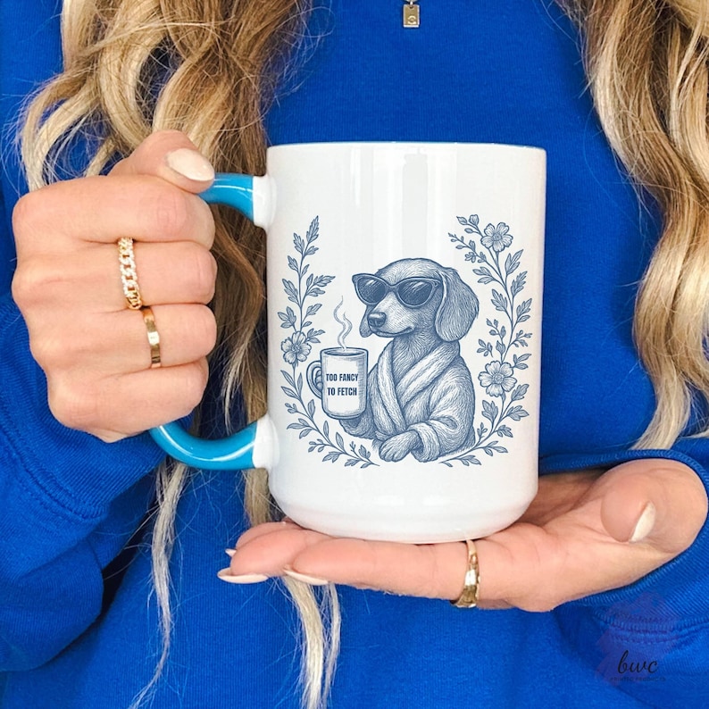 May include: White ceramic mug with a blue handle and rim. The mug features a blue illustration of a dachshund wearing sunglasses and a robe, holding a coffee cup. The text on the cup reads "TOO FANCY TO FETCH".