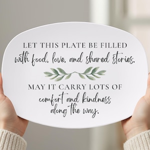 May include: White oval serving plate with black script text and green leaf accents. The text reads: "LET THIS PLATE BE FILLED with food, love, and shared stories. MAY IT CARRY LOTS OF comfort and kindness along the way."