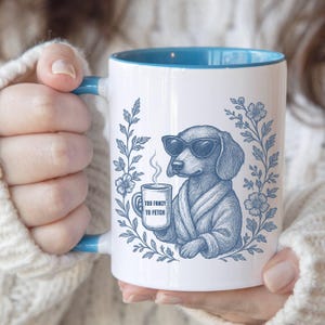 May include: White ceramic mug with a blue interior and handle. The mug features a blue illustration of a dog wearing sunglasses and a robe, holding a coffee cup. Text on the cup reads "TOO FANCY TO FETCH". Floral design surrounds the dog.