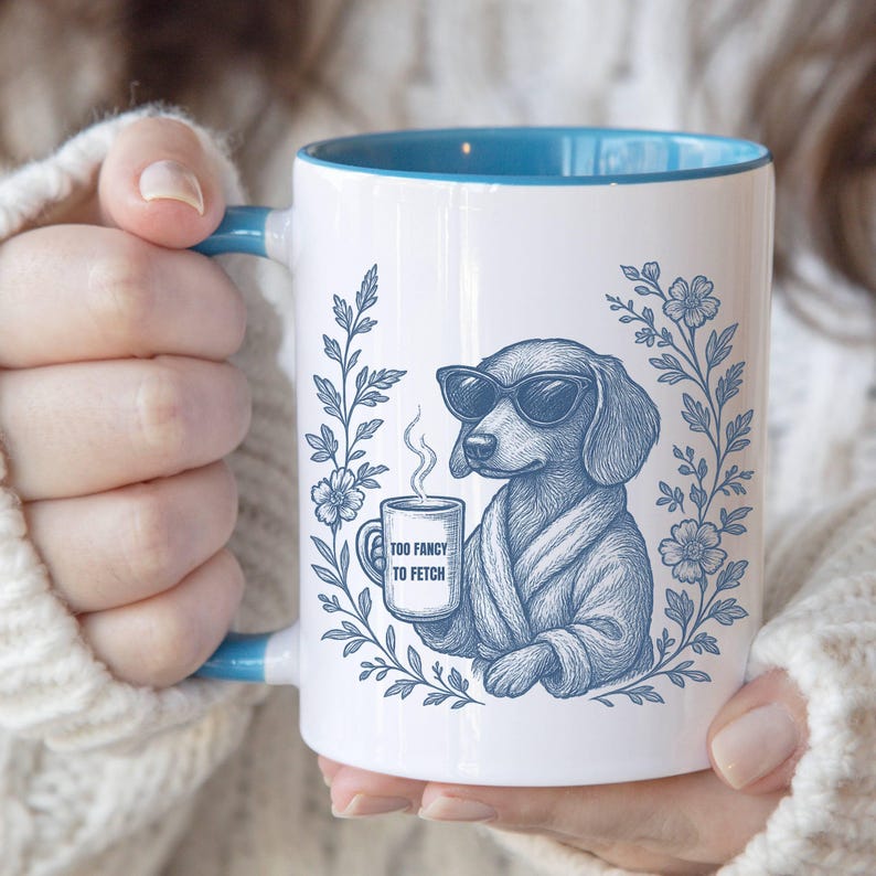 May include: White ceramic mug with a blue interior and handle. The mug features a blue illustration of a dog wearing sunglasses and a robe, holding a coffee cup. Text on the cup reads "TOO FANCY TO FETCH". Floral design surrounds the dog.