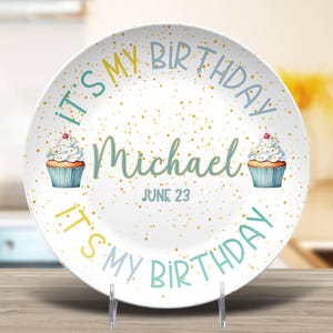 May include: White ceramic birthday plate with gold speckles, the text "IT'S MY BIRTHDAY" in teal and yellow, and the name "Michael" in teal. Two cupcakes with cherries are on the plate. The date "JUNE 23" is also on the plate.