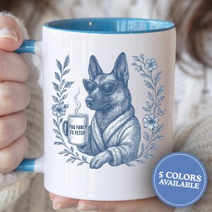 May include: White ceramic mug with a blue interior and handle, featuring a blue illustration of a dog wearing sunglasses and a robe, holding a mug with the text "TOO FANCY TO FETCH". A floral wreath surrounds the dog. The mug is held by a person.