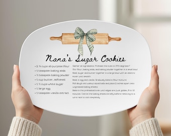May include: A white oval ceramic platter featuring a brown wooden rolling pin illustration with a green and white checkered bow. The text reads Nana's Sugar Cookies followed by a list of ingredients and baking instructions in black script.