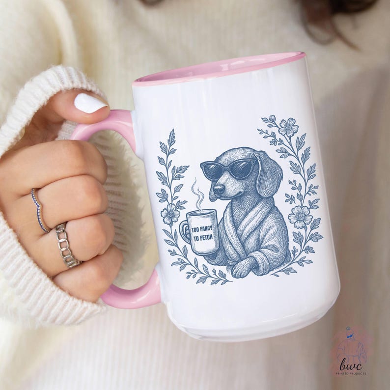 May include: White ceramic mug with a pink interior and handle. The mug features a blue illustration of a dachshund wearing sunglasses and a robe, holding a coffee cup. The cup reads "Too Fancy to Fetch".