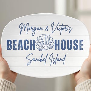 May include: White, oval-shaped sign with blue text that reads "Morgan & Victor's BEACH HOUSE Sanibel Island." A seashell graphic is between "BEACH" and "HOUSE." The sign has a wood-grain background.