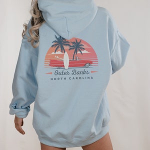 May include: A light blue hooded sweatshirt with a graphic print of a red car driving on a beach with palm trees and a surfboard. The text "Outer Banks" and "NORTH CAROLINA" is printed below the graphic.