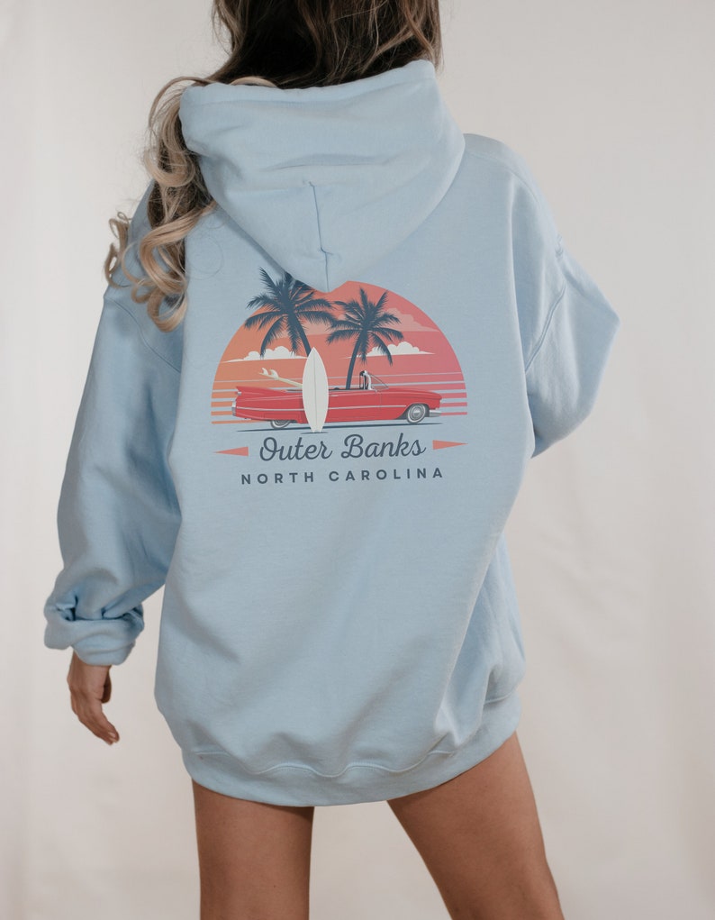 May include: A light blue hooded sweatshirt with a graphic print of a red car driving on a beach with palm trees and a surfboard. The text "Outer Banks" and "NORTH CAROLINA" is printed below the graphic.