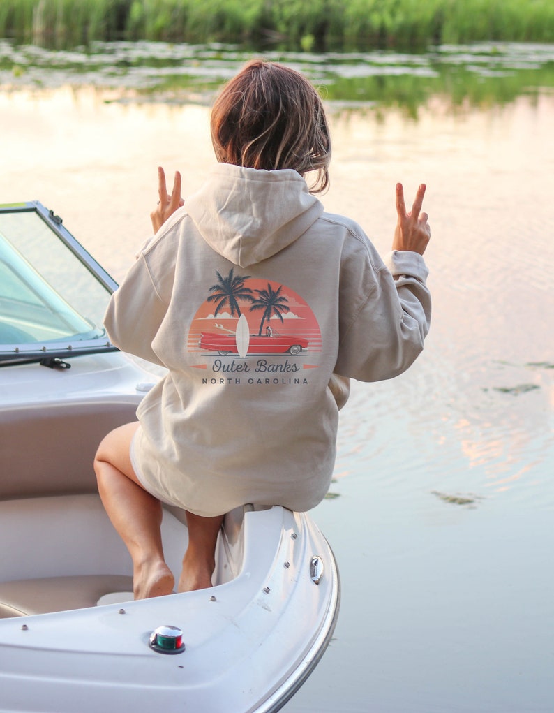 May include: A person wearing a beige hoodie with a graphic of palm trees, a surfboard, and a red car with the text "Outer Banks North Carolina".