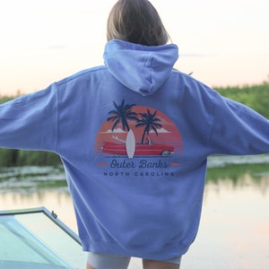 May include: A light blue hooded sweatshirt with a graphic print of a red car driving on a beach with palm trees and a surfboard. The text "Outer Banks North Carolina" is printed below the graphic.