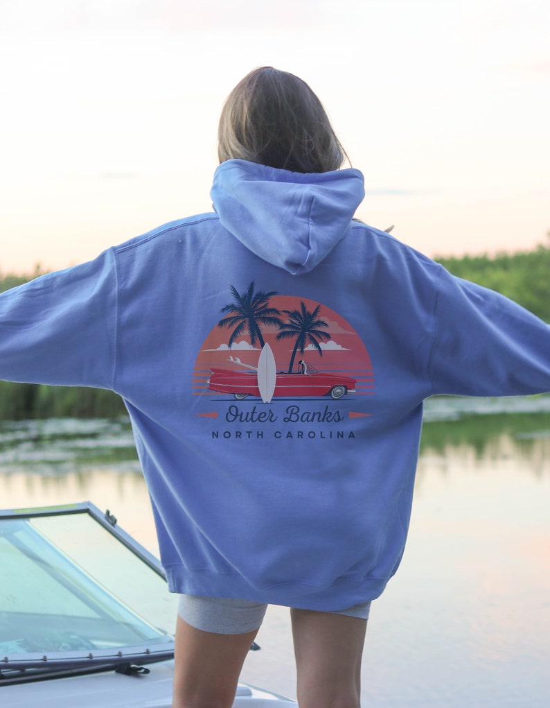 May include: A light blue hooded sweatshirt with a graphic print of a red car driving on a beach with palm trees and a surfboard. The text "Outer Banks North Carolina" is printed below the graphic.