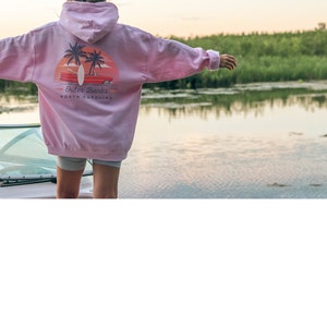 May include: A pink hoodie with a graphic print of a sunset, palm trees, and a surfboard with the text "Outer Banks, North Carolina".