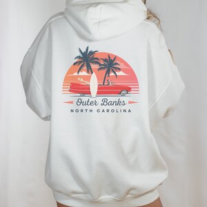 May include: White hooded sweatshirt with a graphic print of a red car, surfboard, and palm trees against a sunset background. The text "Outer Banks North Carolina" is printed below the graphic.